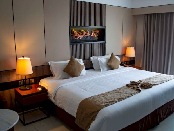 The image shows a hotel room with a large bed, decorative pillows, bedside lamps, and a small table, creating a cozy atmosphere.
