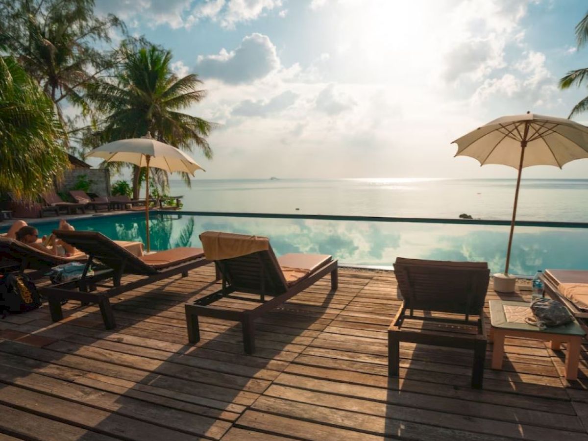 A serene tropical poolside scene with palm trees, lounge chairs, umbrellas, and a view of the ocean under a partly cloudy sky.