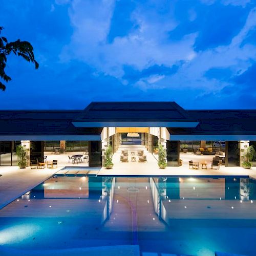 The image shows a modern building with a large illuminated pool in the foreground, surrounded by stylish outdoor seating areas.