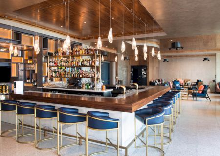 The image shows a modern bar with a wooden countertop, high chairs, and pendant lights, creating a cozy atmosphere.
