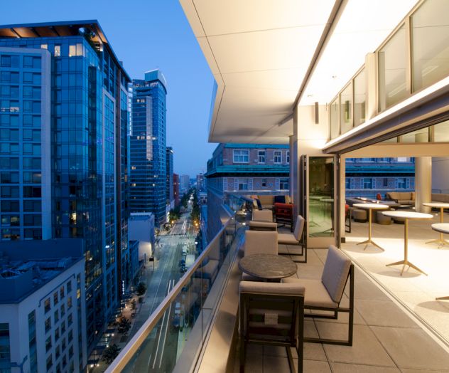 Modern rooftop patio with seating overlooks a cityscape at dusk, featuring tall buildings and a street below.