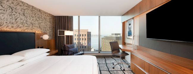 A modern hotel room with a king-size bed, large TV, desk, chair, and a window with a city view.