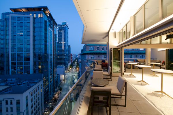 A modern rooftop terrace at dusk with seating, overlooking a city street and high-rise buildings under a twilight sky.