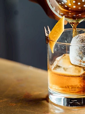 A drink is being poured into a glass with ice and a citrus twist garnish, against a dark background.