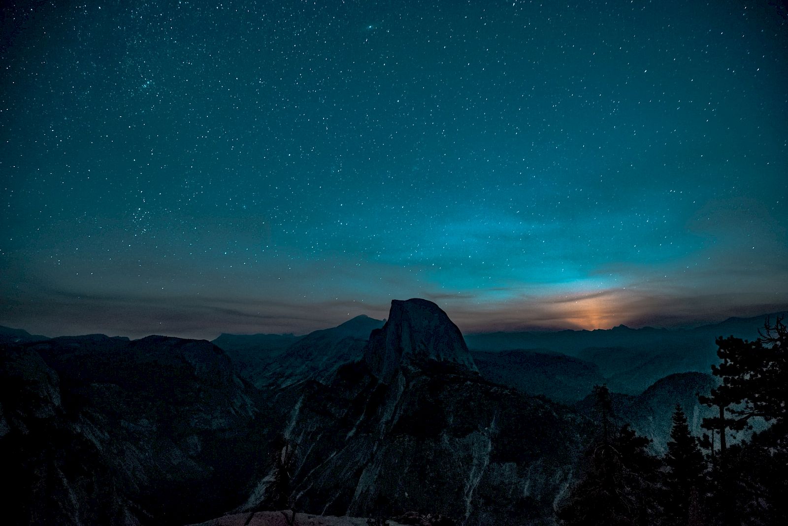 A starry night sky over a mountainous landscape with a prominent peak and distant horizon glow, under a cool blue atmosphere.