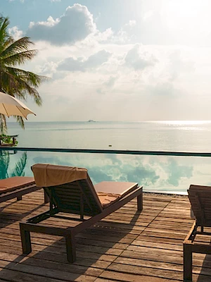 A serene beach scene with lounge chairs, umbrellas, and palm trees by a pool overlooking a calm sea under a partly cloudy sky.