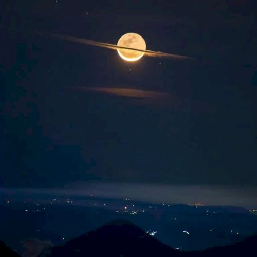 A full moon is surrounded by clouds, resembling Saturn's rings, over a landscape with distant city lights visible in the night.