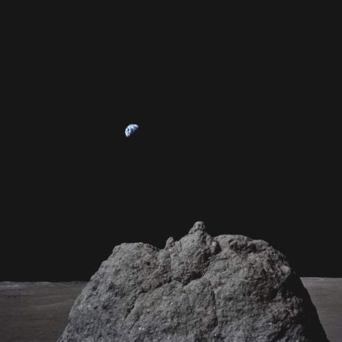 A lunar surface with a large rock in the foreground and Earth visible in the dark sky above.