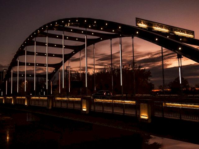 A lit arched bridge over a dark river at dusk, with strings of lights and a stadium-style sign at the top.