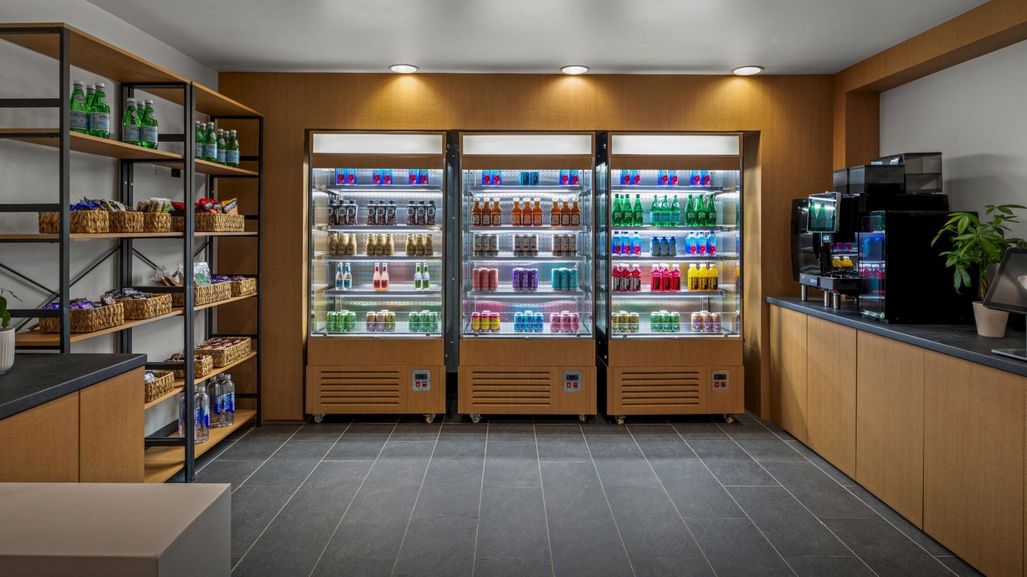 A small shop with three glass-front fridges filled with colorful drinks, shelves of snacks, and a counter with a coffee machine on the right.