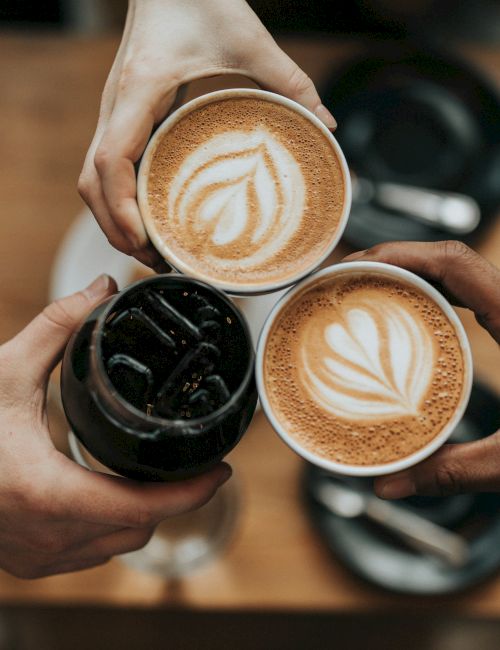 Three hands hold up two lattes with latte art and one iced coffee over a table, suggesting a coffee gathering or toast.