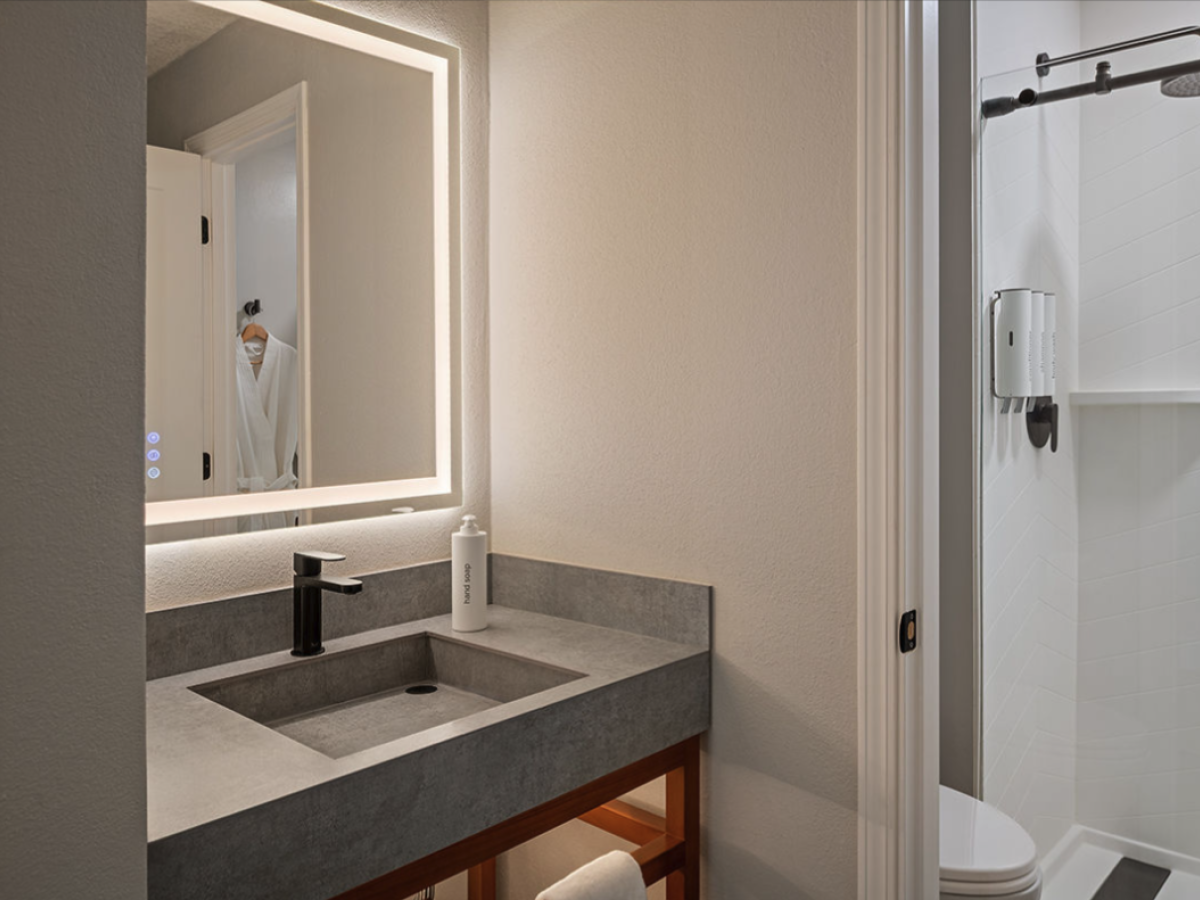 A modern hotel bathroom with a backlit mirror, gray concrete countertop, black faucet, sink, wooden shelf, and a separate glass-enclosed shower.