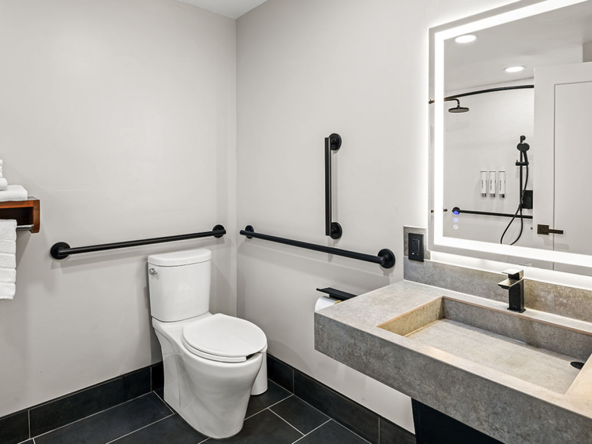 Modern bathroom with a toilet, grab bars, concrete-style sink, and a large illuminated mirror and shower controls.