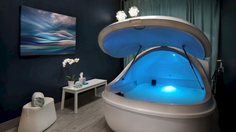 A room with a sensory deprivation tank, ambient blue lighting, a decorative table, wall art, orchid, and a small decorative item.