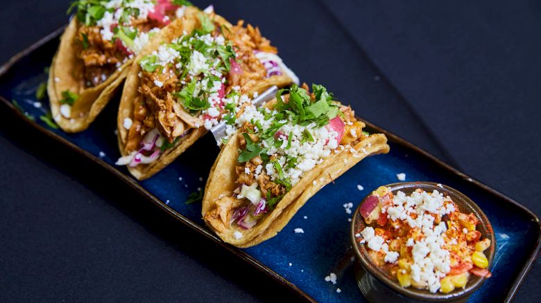 The image shows delicious tacos filled with meat, cilantro, and cheese, served with salsa in a bowl on the side, on a blue plate.