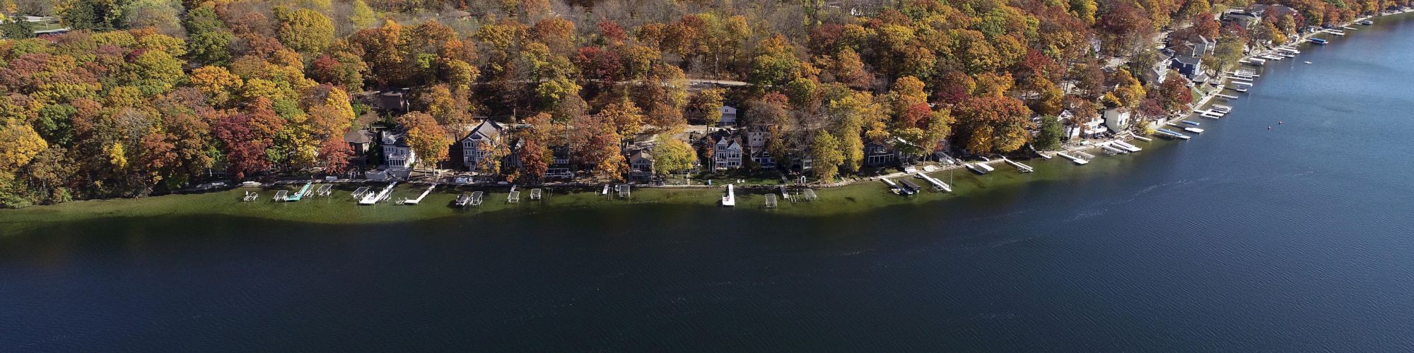 Aerial view of a lake surrounded by autumn trees, docks, and houses, with a clear, expansive sky in the background, showcasing fall colors.