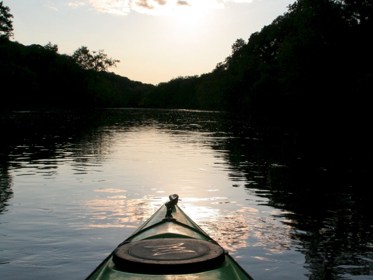 A kayak on calm water at sunset, with trees lining both sides and clouds in the sky, creating a serene and picturesque scene.
