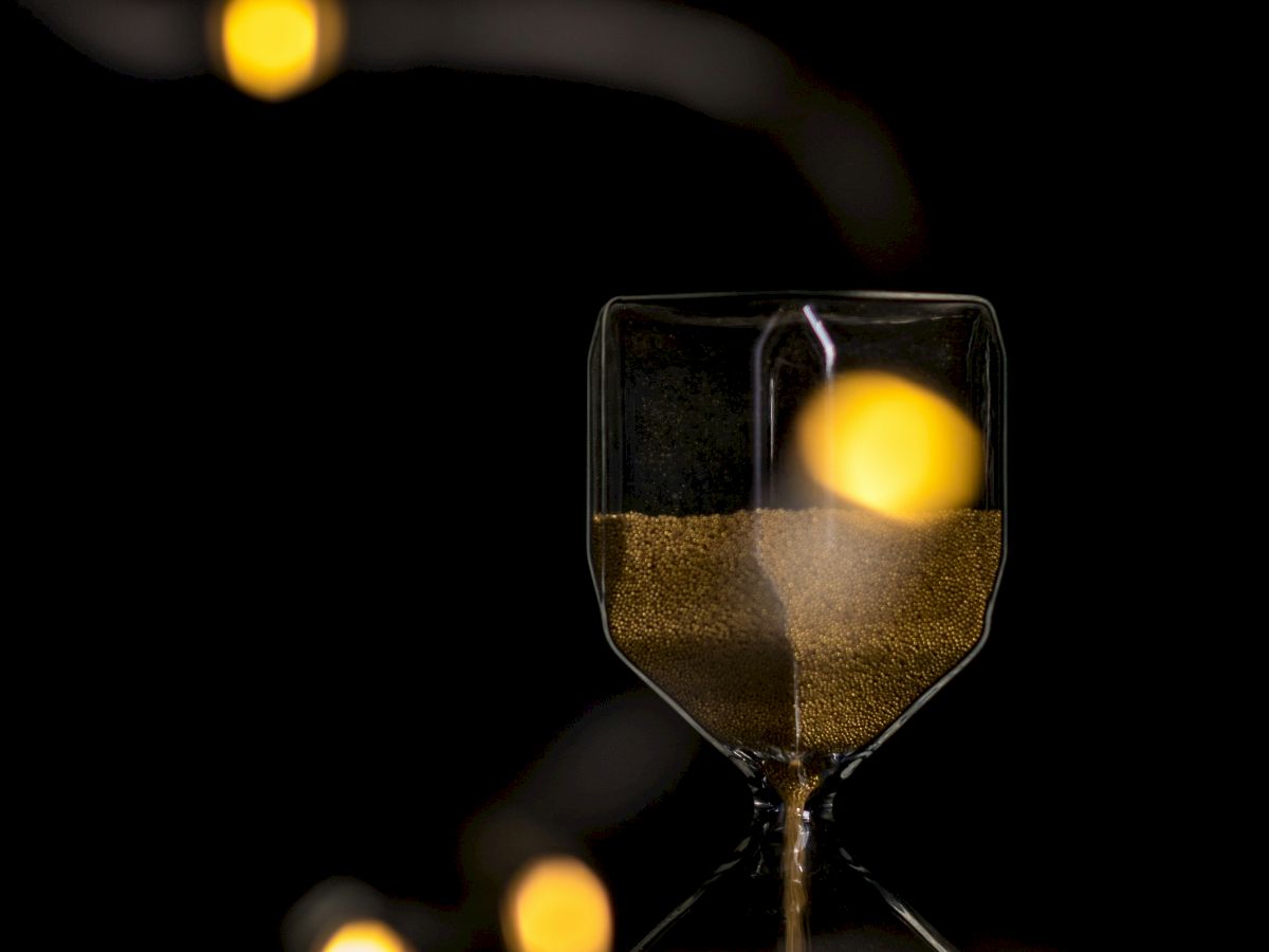 An hourglass with cascading sand is set against a dark backdrop, adorned with soft, golden bokeh lights trailing diagonally.