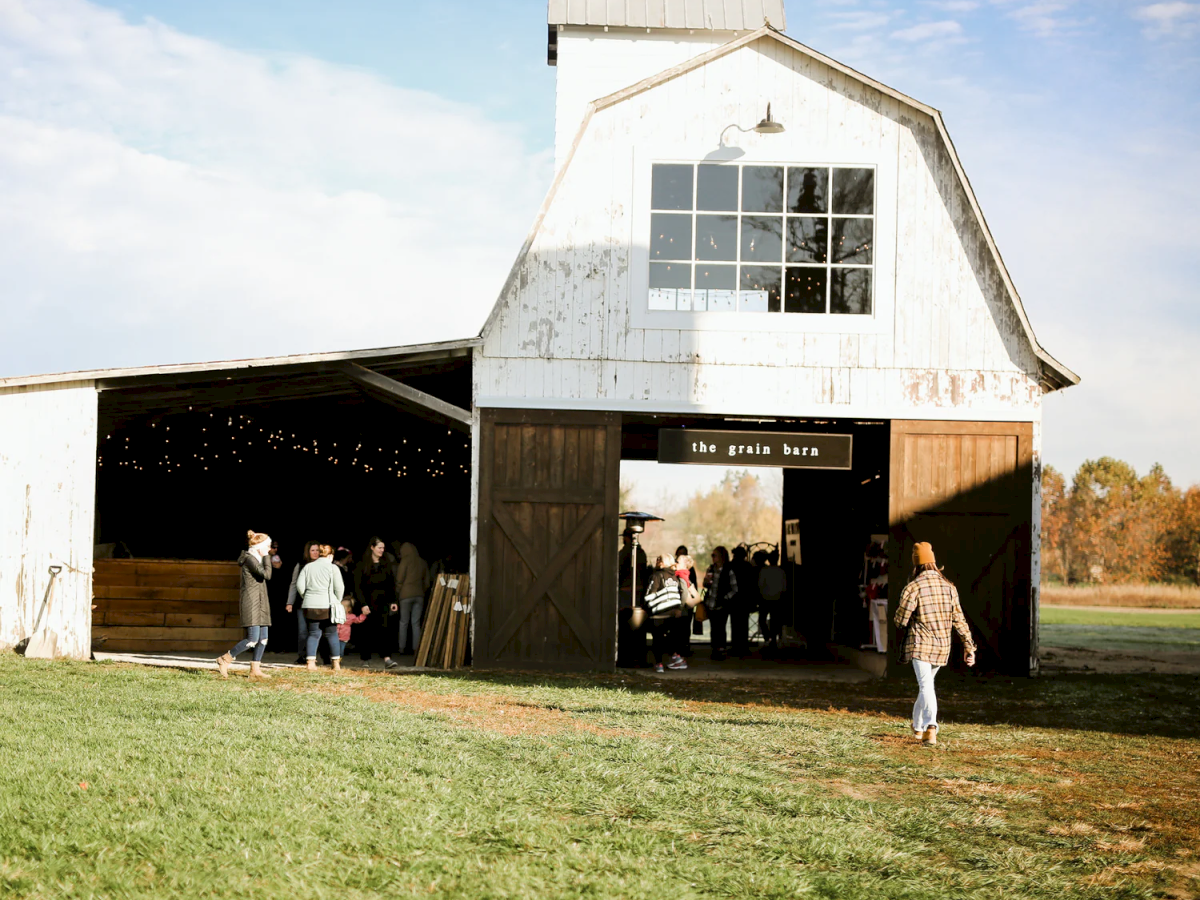 A white barn with large doors open, people entering and exiting, surrounded by green grass and trees in the background.