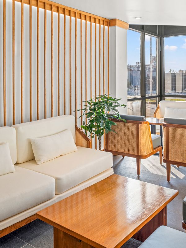 A stylish, sunlit living room with cream sofas, wooden coffee table, striped wood wall, and a bright windowed seating area overlooking the city, cozy and modern.