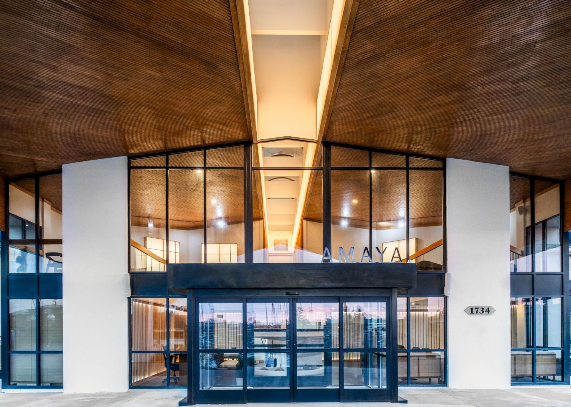Modern building entrance with glass doors, an illuminated indoor corridor, wood ceiling, and an &ldquo;AMAYA&rdquo; sign with number 1734.