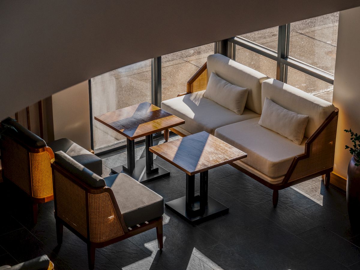 Sunlit terrace with a beige lounge sofa, cushioned chairs, and wooden coffee tables by a glass railing.