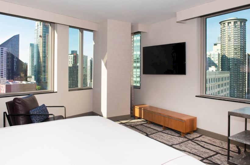 A modern hotel room with large windows showing a cityscape, wall-mounted TV, bed, chair, and a small table.