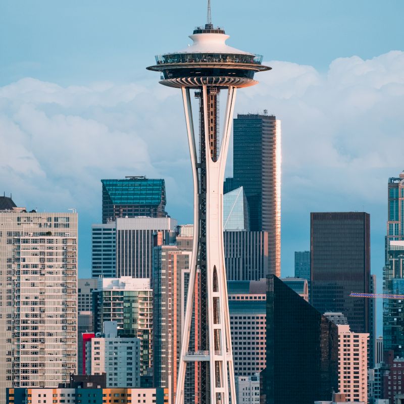The image shows the Space Needle set against a city skyline with tall buildings and a cloudy sky backdrop.