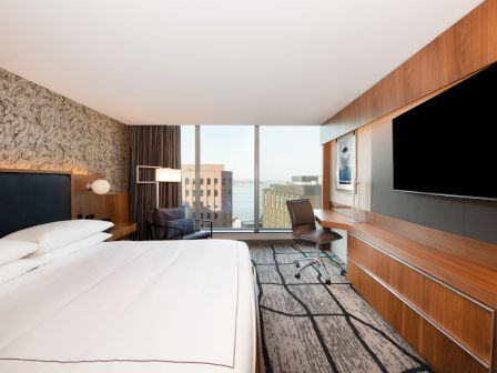 The image shows a modern hotel room with a large bed, a wall-mounted TV, a desk and chair, and a view of buildings through a window.