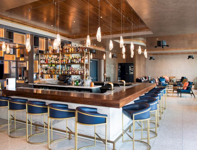 The image shows a modern bar with a wooden counter, blue stools, hanging lights, and shelves stocked with bottles, in a spacious setting.
