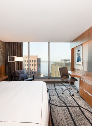 A modern hotel room with a king-sized bed, large TV, desk, chair, and a window with a city view.