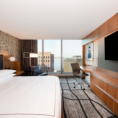 A modern hotel room features a bed, desk, large TV, and window with a city view. Warm tones and sleek d&eacute;cor create a cozy atmosphere.
