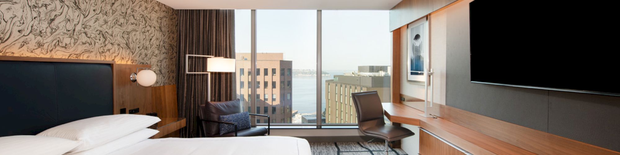 The image shows a modern hotel room with a large bed, wall-mounted TV, desk, and a window offering a cityscape view.